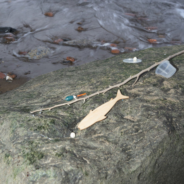 A wooden fish-shaped hand reel and fishing line with a bobber rest on a rock by a stream.