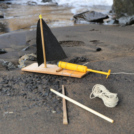 A DIY toy sailboat with a black sail and yellow propeller motor on a sandy beach, featuring wooden sticks and twine.