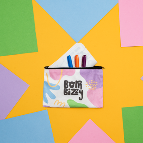 A zippered pouch labeled "born bizzy" sits on a yellow background surrounded by colorful sheets of paper.