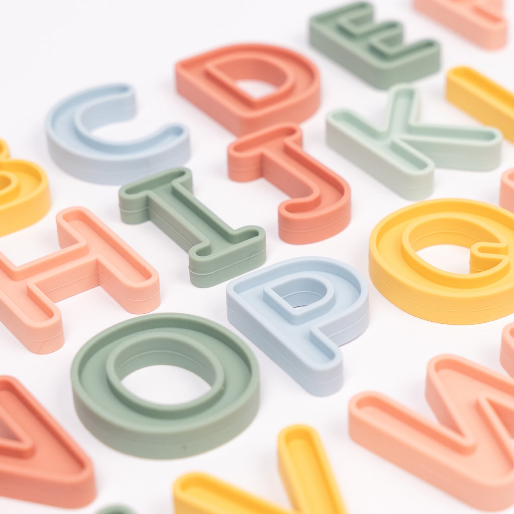 Alphabet letters in pastel colors are arranged randomly. The letters feature hues of green, pink, light blue, and yellow, creating a playful and cheerful tone.