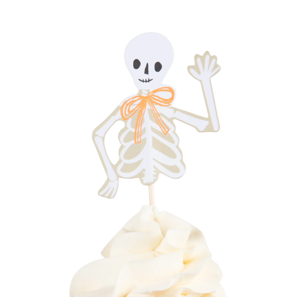 Paper skeleton with an orange bow waves on top of a creamy, white cupcake. Set against a white background, it conveys a whimsical Halloween vibe.