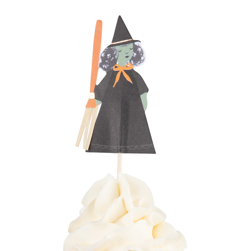 Paper witch figure with green skin and black hat on a stick, adorned with an orange bow and broom, stands atop a swirl of white frosting. Halloween theme.