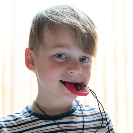 A young boy smiling while chewing on a red circular dinosaur chew pendant necklace.