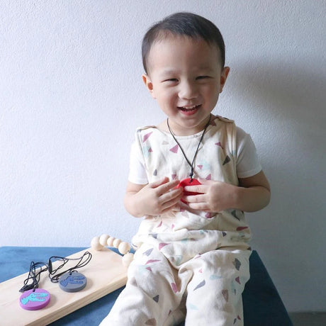 Smiling child wearing a red dino sensory pendant necklace with extra colorful pendants displayed nearby.