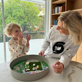Woman and child playing together with a Jellystone Designs fairy garden sensory kit near a large window.