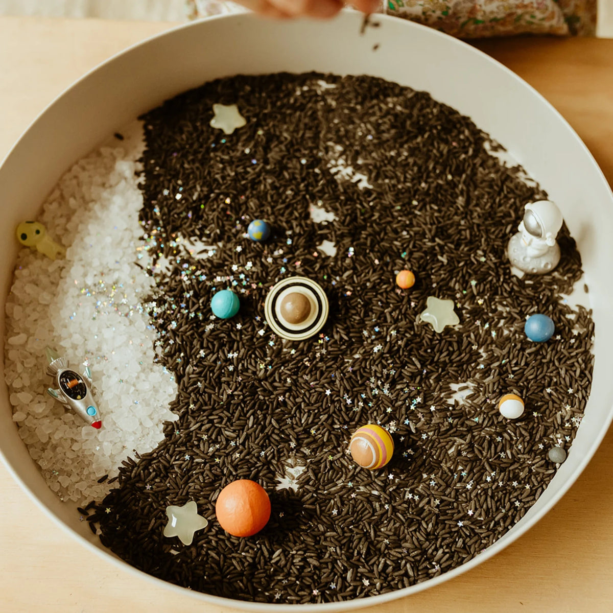 Galaxy sensory play tray filled with dark rice, miniature planets, stars, an astronaut, and a rocket.