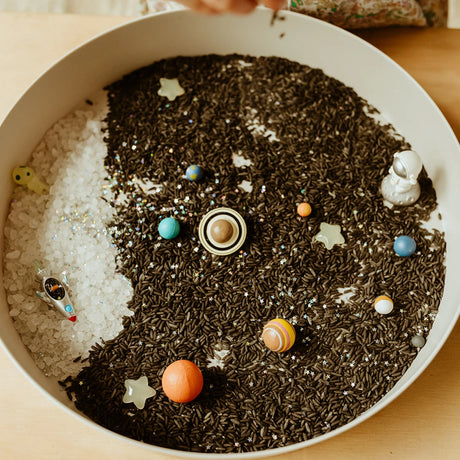 Galaxy sensory play tray filled with dark rice, miniature planets, stars, an astronaut, and a rocket.