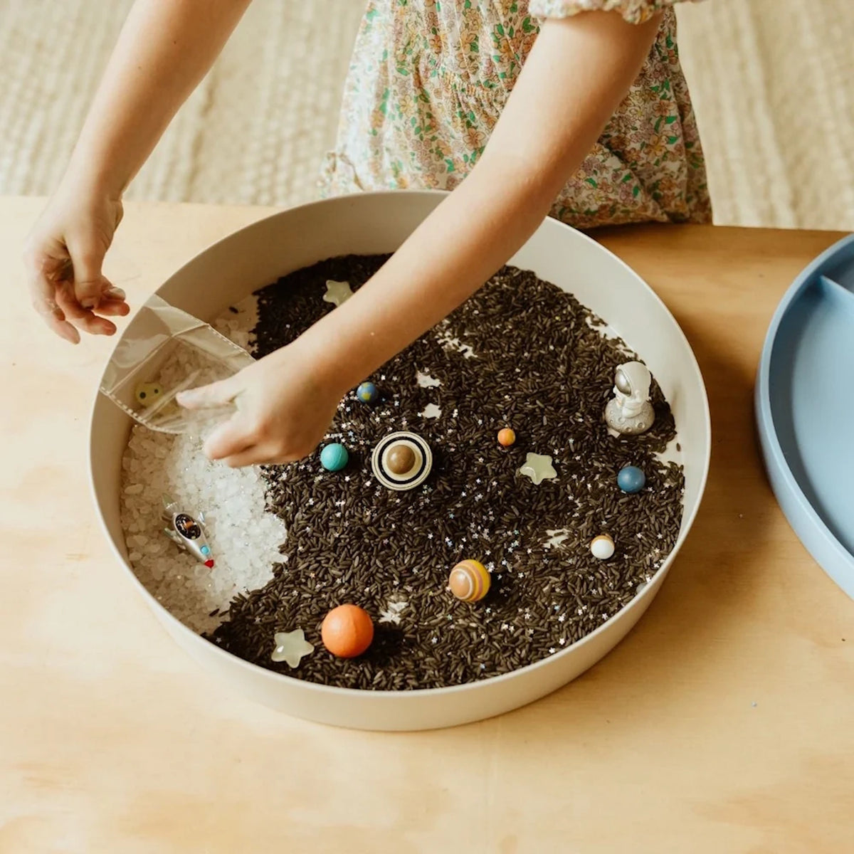 Child pouring trinkets into a round galaxy sensory tray filled with black rice, white rocks, and planets.