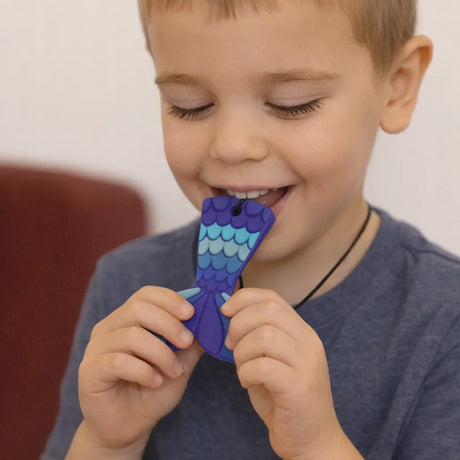 Young boy happily chewing on a purple and blue silicone mermaid tail sensory pendant necklace.