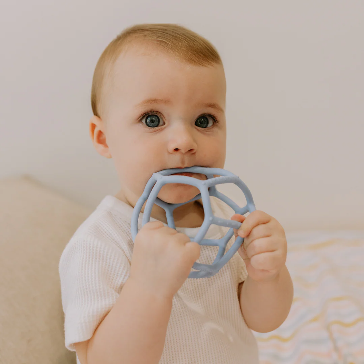 A baby with blue eyes wearing a white shirt chews on a blue, spherical toy. The background is soft and neutral, conveying a calm atmosphere.