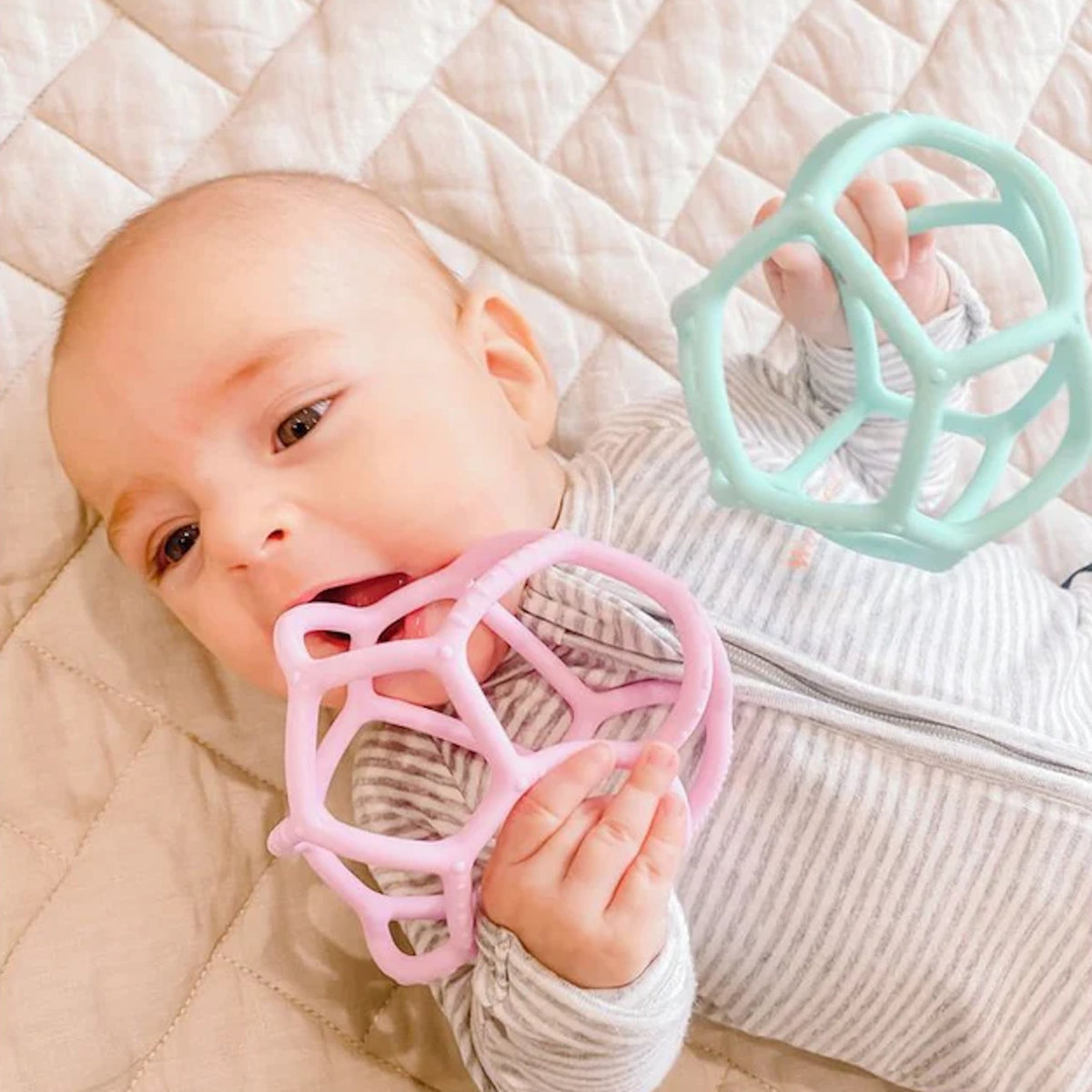 Baby lying down, chewing a pink silicone sensory ball while holding a mint green sensory teether ball.