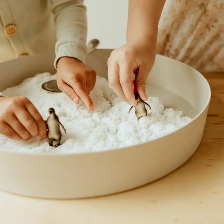 Children's hands playing with toy penguins in a round tray filled with white sensory snow.