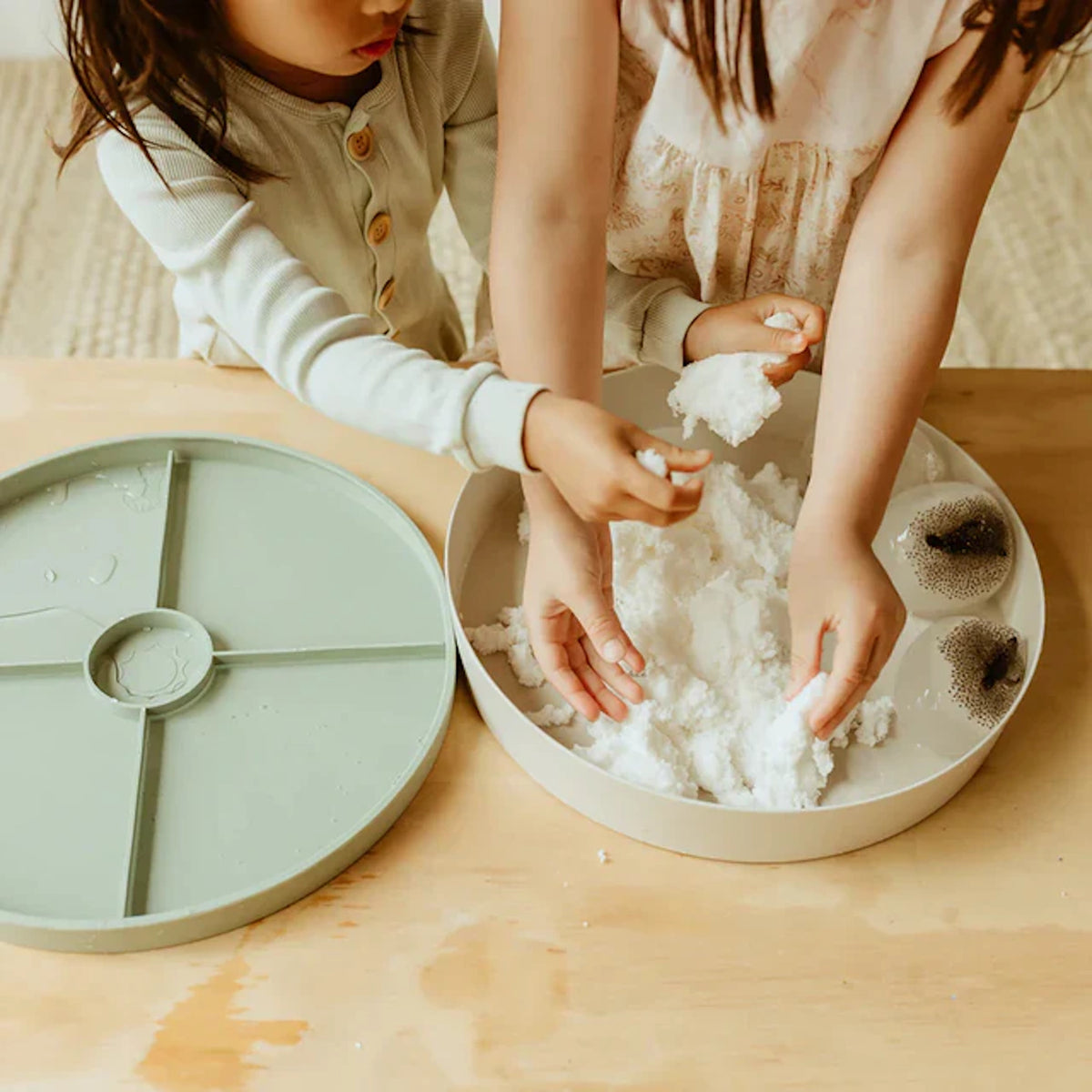 Two children playing with white sensory snow inside a circular tray on a wooden table.