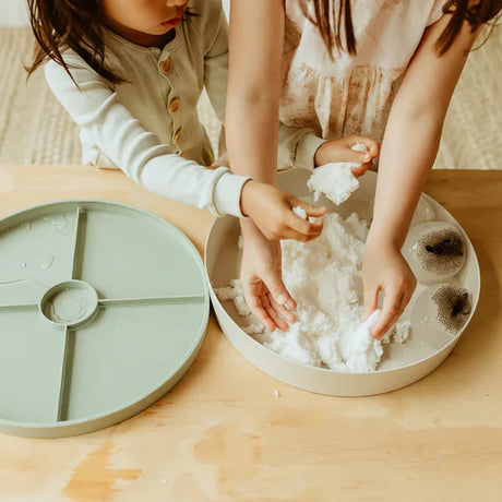 Two children playing with white sensory snow inside a circular tray on a wooden table.
