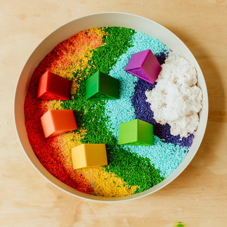 Round sensory tray with rainbow-colored rice, colorful toy houses, and white sensory snow.