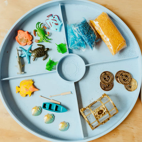 Light blue divided play tray arranged with sea creature toys, colored rice, toy coins, and a treasure chest.