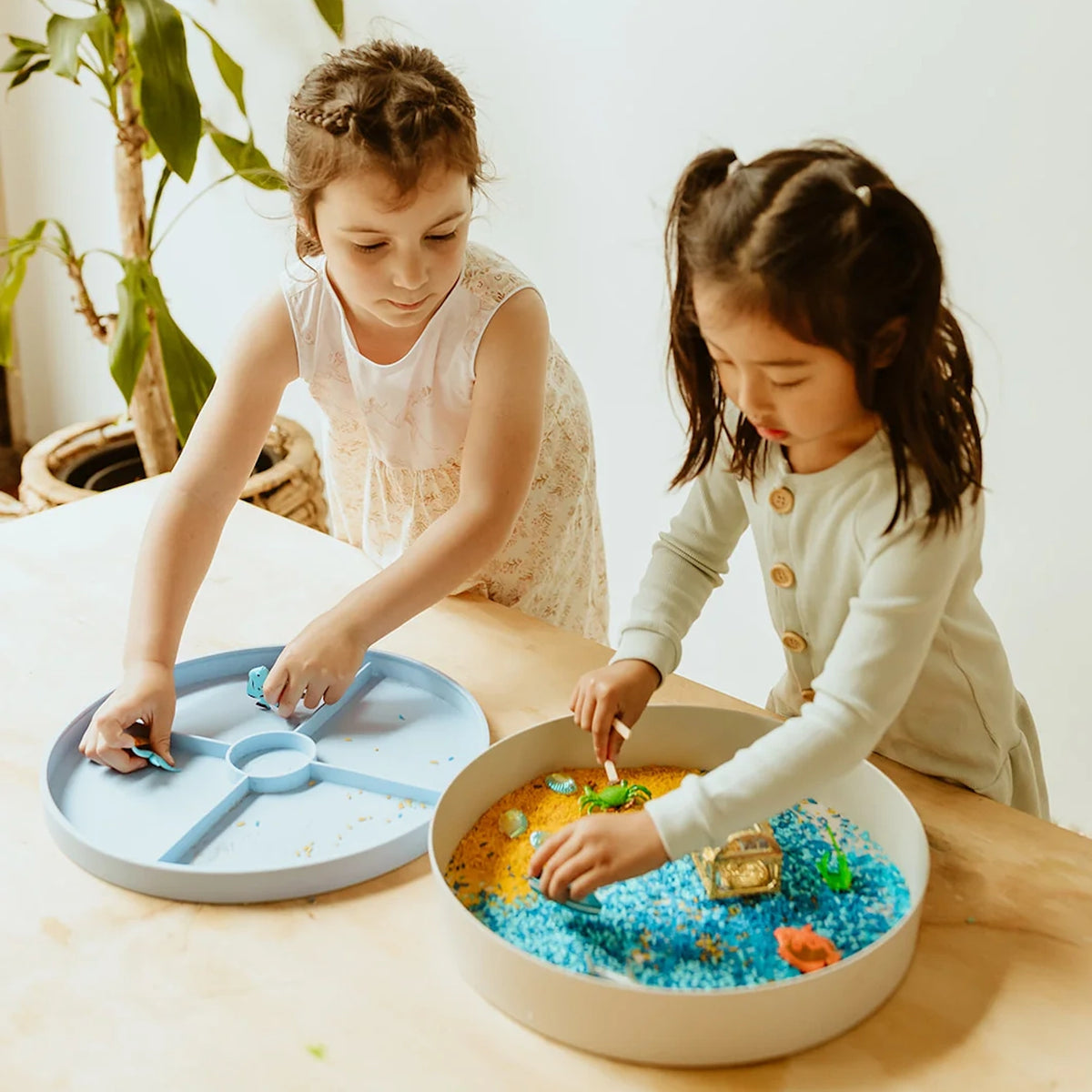 Two young girls playing with an ocean-themed sensory play tray filled with colorful rice and sea toys.