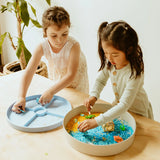 Two young girls playing with an ocean-themed sensory play tray filled with colorful rice and sea toys.