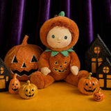 Plush doll in a jack-o'-lantern suit sitting among small pumpkins and haunted paper houses.