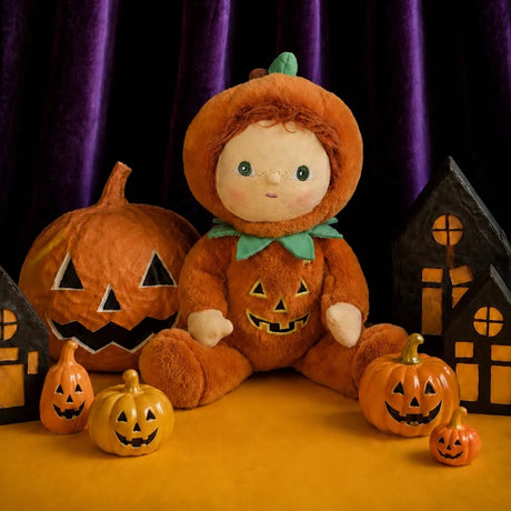 Plush doll in a jack-o'-lantern suit sitting among small pumpkins and haunted paper houses.