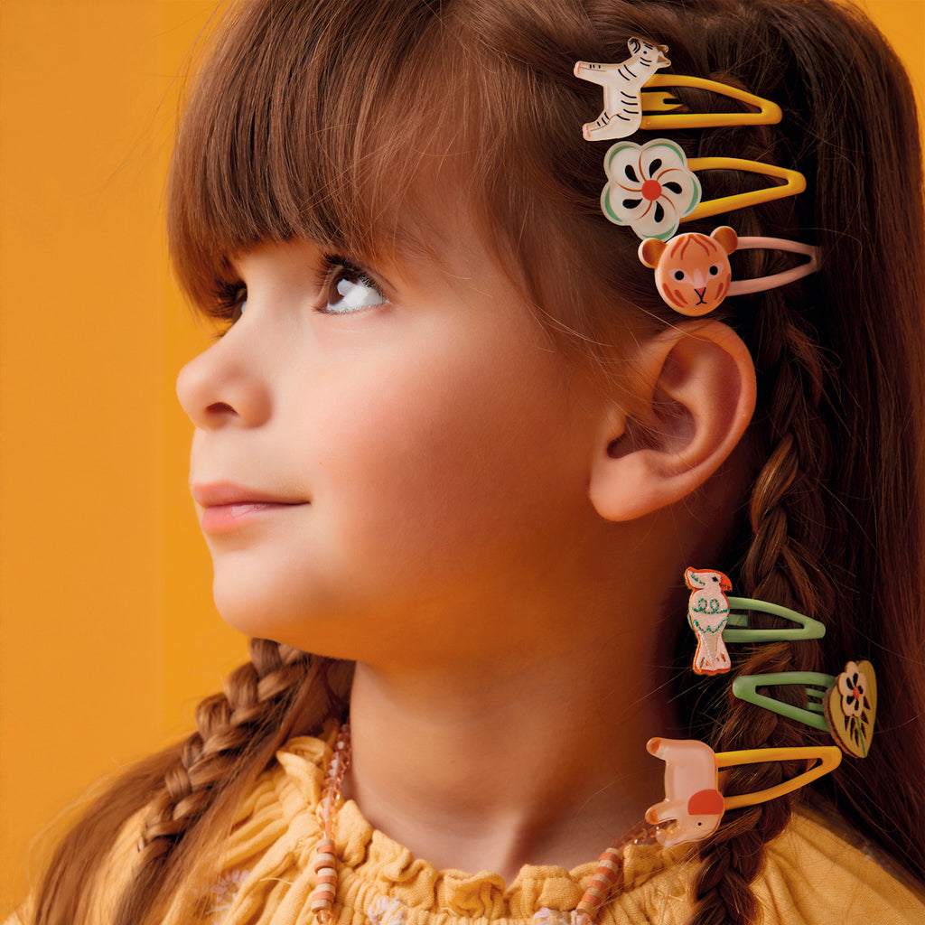 A young girl with braided hair looks to the side, wearing colorful clips shaped like animals and flowers. The cheerful orange background matches her yellow outfit.