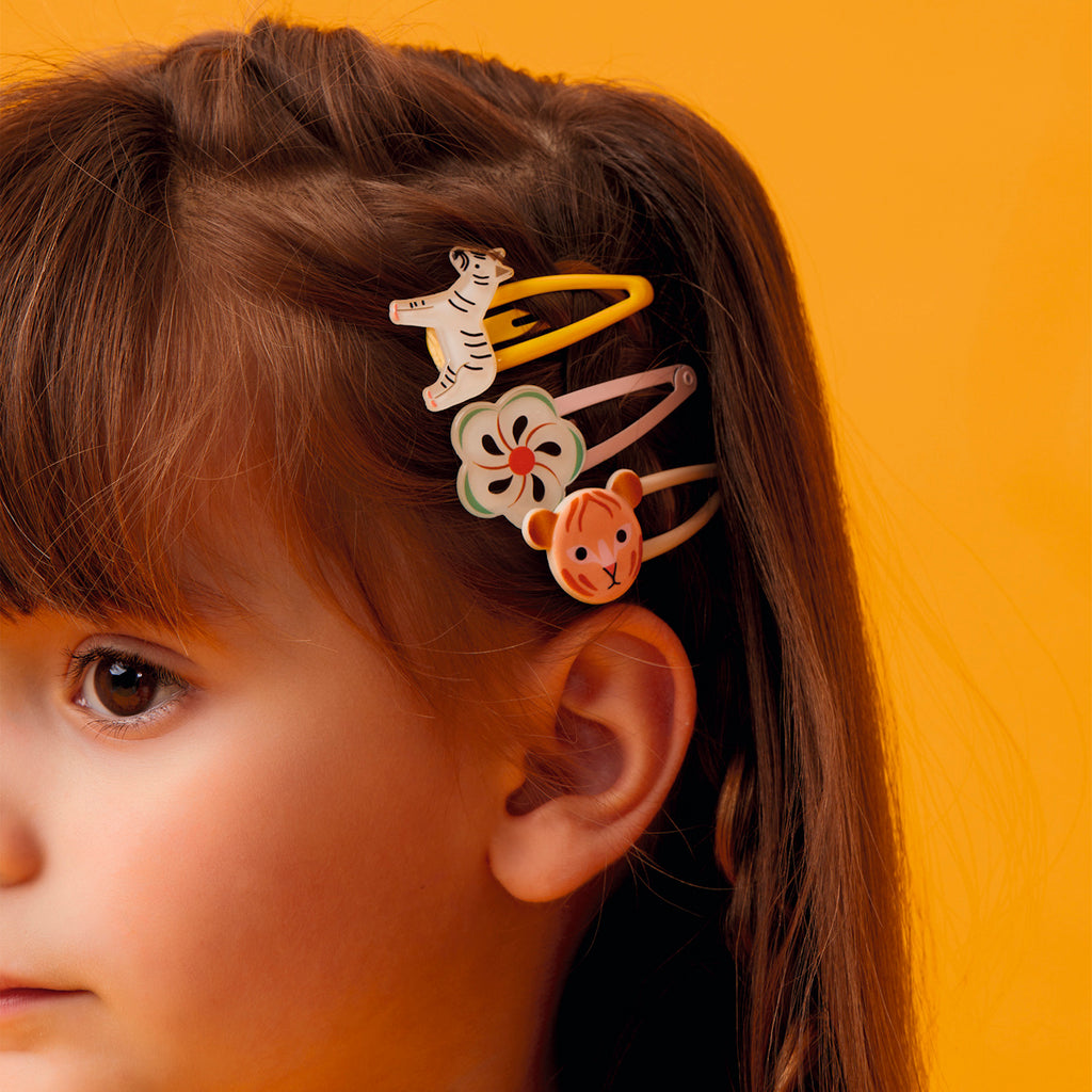 Close-up of a child's hair adorned with colorful clips shaped like a white tiger, a flower, and an orange bear. Background is vibrant orange.