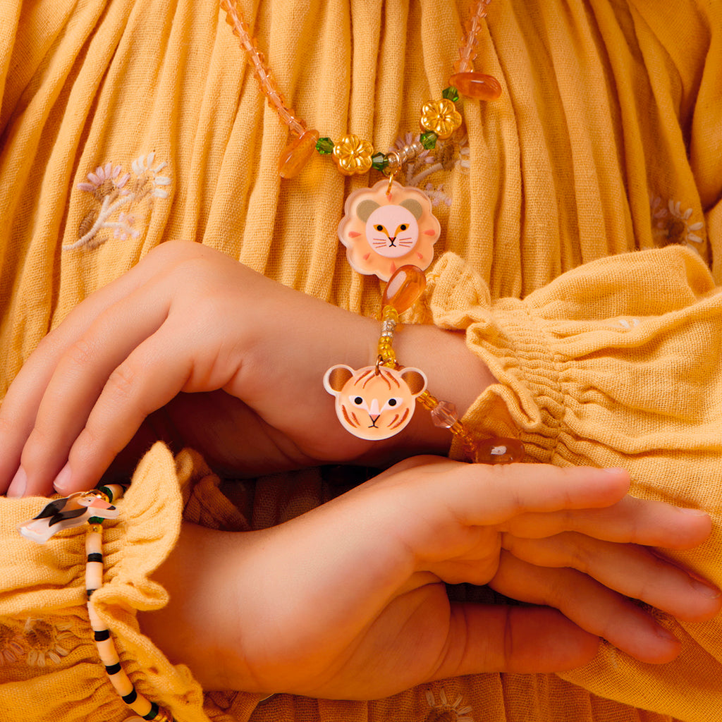 A young girl in a yellow dress gazes down at her necklace and bracelet with lion faces. She has floral hair clips and the background is a matching yellow.
