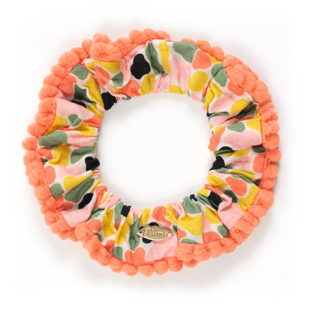 Colorful fabric scrunchie with a floral pattern in pink, green, yellow, and black. It features orange pom-pom trim and a small gold tag. Fun and vibrant.