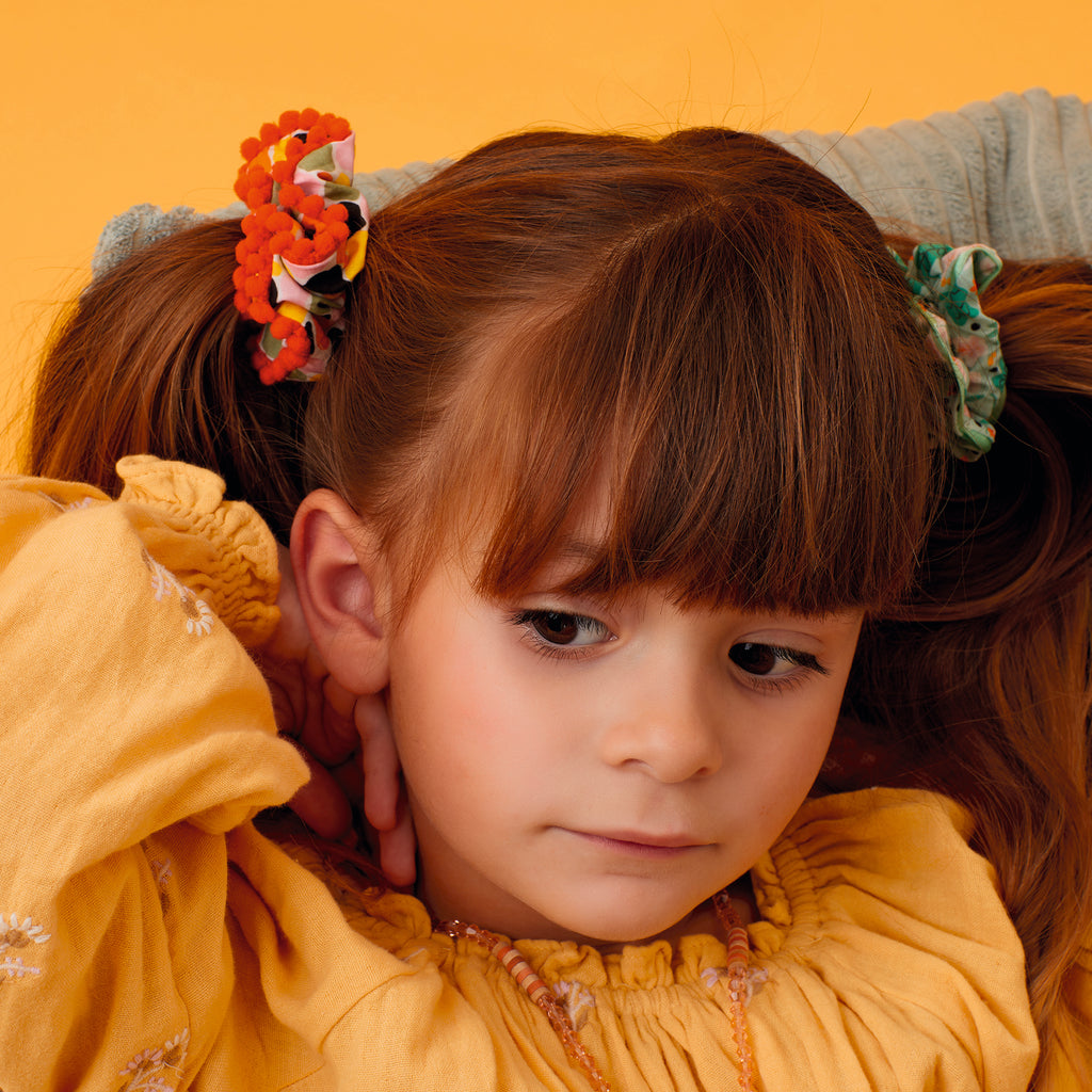 A young girl with brown hair and a thoughtful expression, wearing a yellow dress. Her hair is styled in ponytails adorned with colorful flower clips. The background is a warm, muted orange.