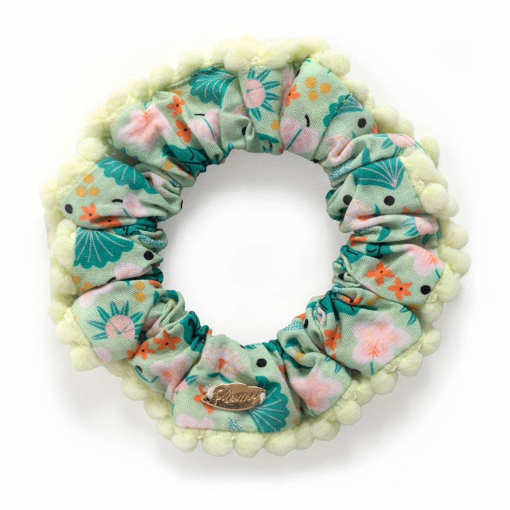 A pastel green scrunchie with floral and abstract patterns in pink and orange. It features a fluffy trim and a small metal tag with text, conveying a playful and stylish tone.