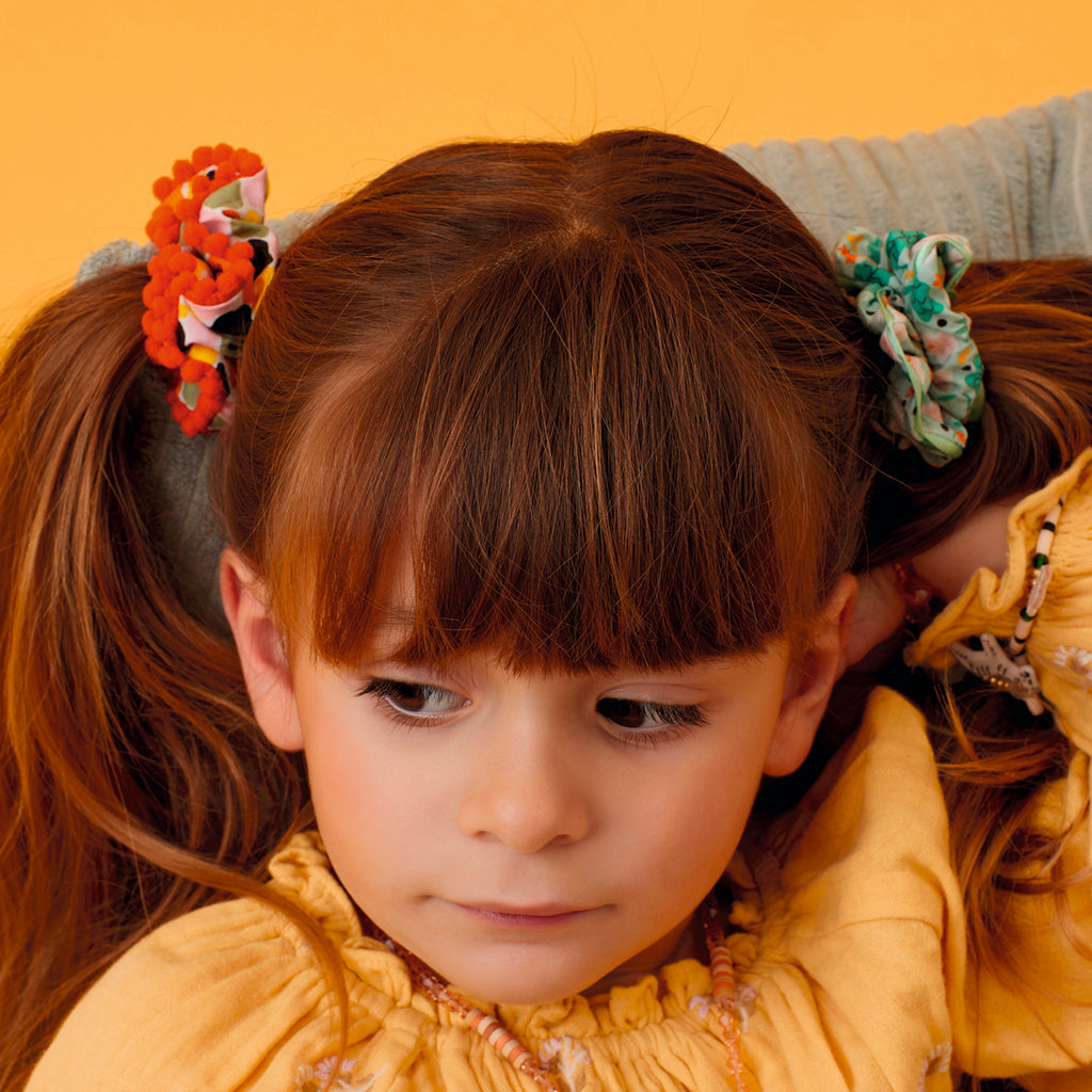 Young girl with brown hair in pigtails adorned with colorful scrunchies, wearing a yellow shirt. She gazes thoughtfully to the side on a warm orange backdrop.