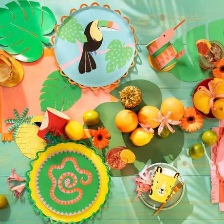 Jungle-themed party table with animal plates, colorful cups, tropical leaves, and fresh citrus fruits.