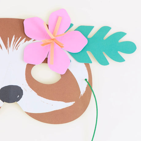 A close-up of a paper sloth party mask with a pink flower and green leaf detail on a white background.