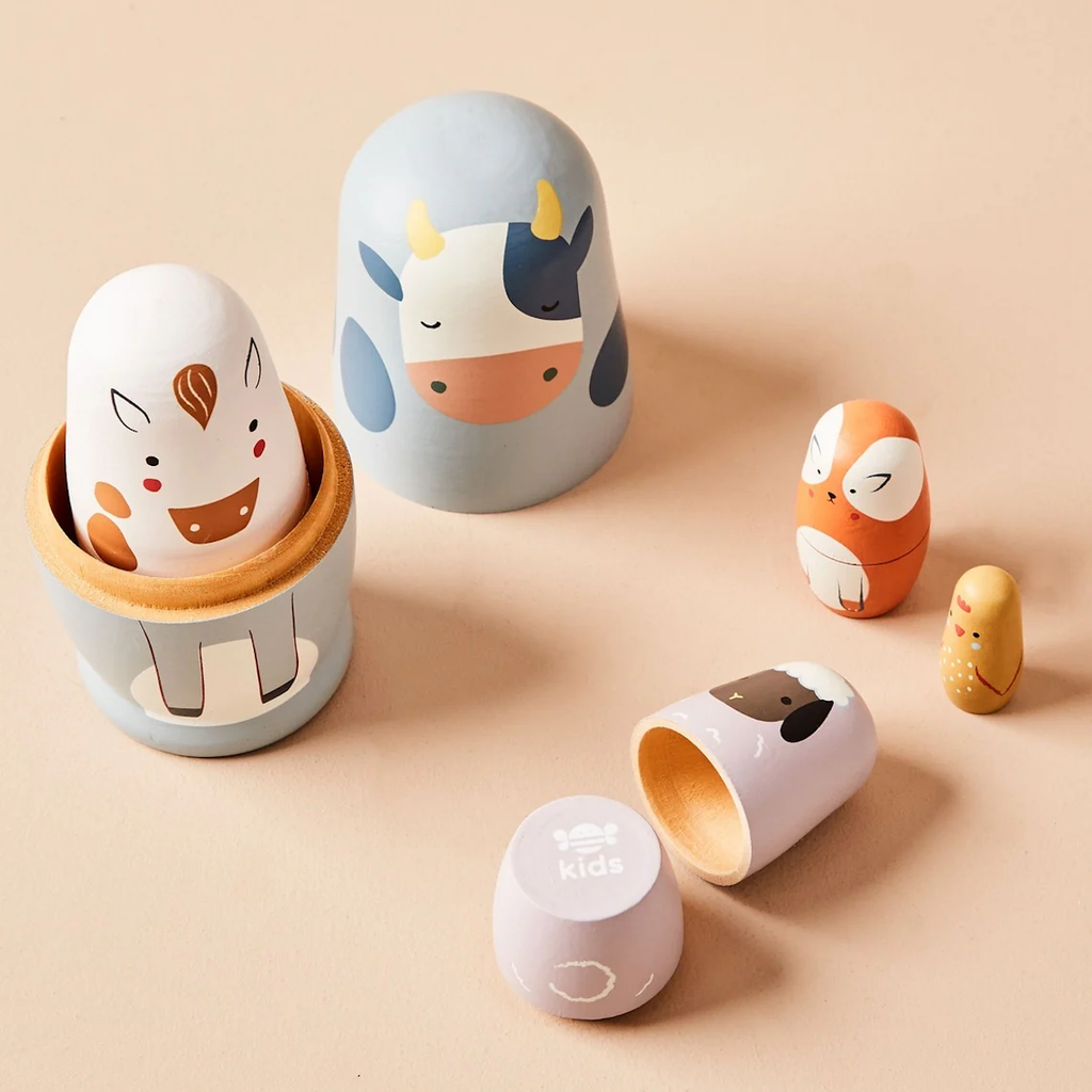 Toy nesting dolls painted as animals on a peach background. Includes a cow, horse, fox, sheep, and a small chick. The mood is playful and cheerful.