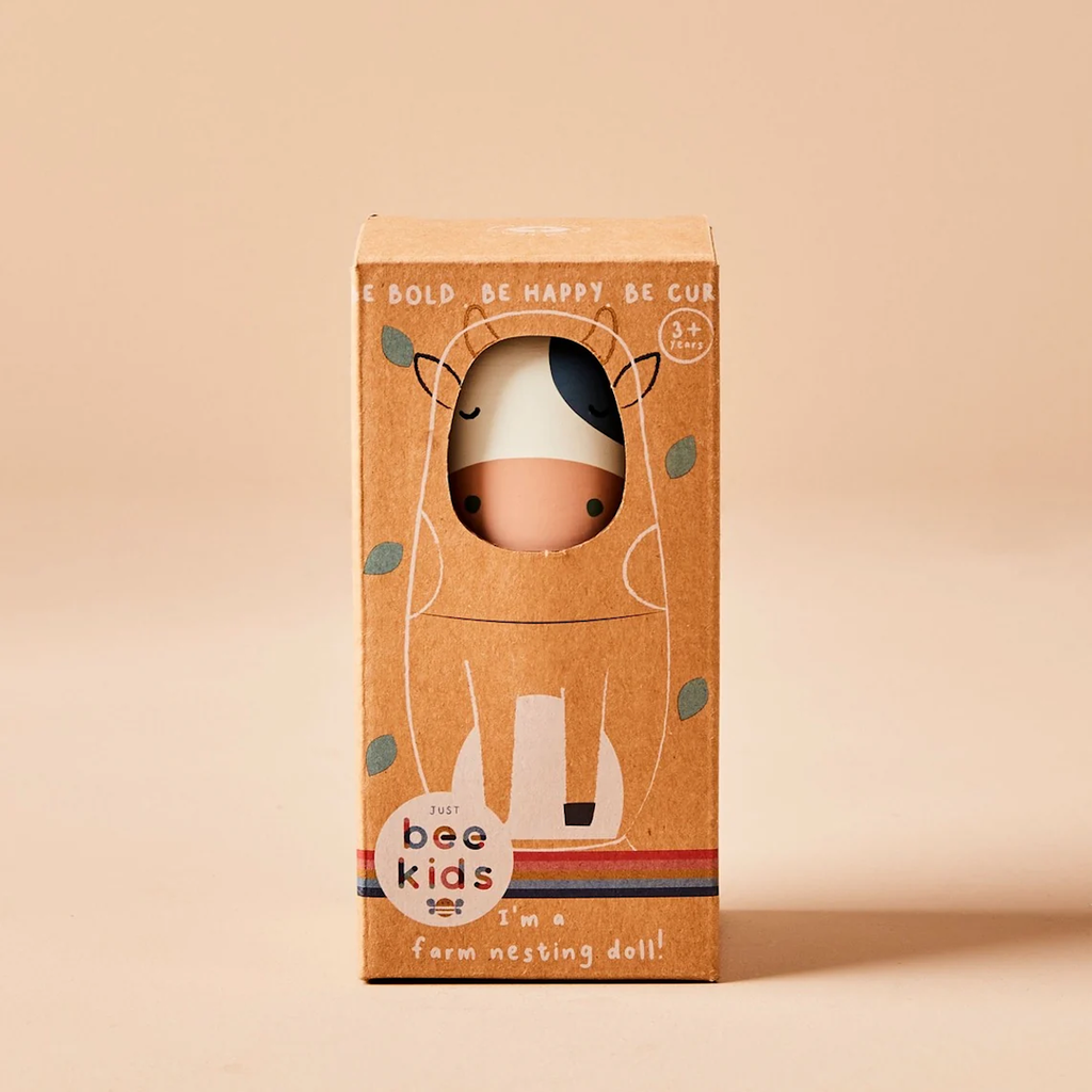 Brown packaging box for a farm-themed nesting doll, featuring a cute cow illustration and rainbow stripes. Text reads "Just bee kids, I'm a farm nesting doll!" The tone is playful and inviting.