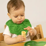 Baby in a white shirt and green leaf-shaped bib eating a banana while sitting in a wooden high chair.