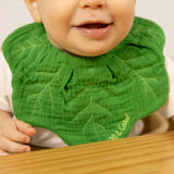 Smiling baby wearing a green leaf-shaped muslin bib with embroidered veins sitting at a wooden table.