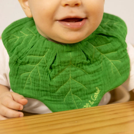 Smiling baby wearing a green leaf-shaped muslin bib with embroidered veins sitting at a wooden table.