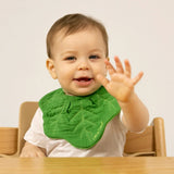 Smiling baby in a high chair wearing a green kale leaf-shaped muslin bib, waving one hand toward the camera.