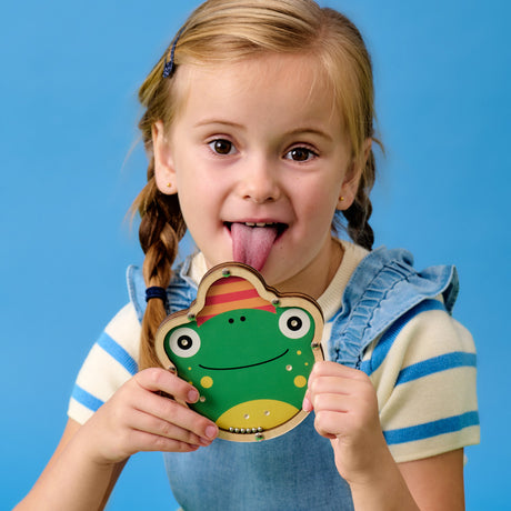 Young girl in blue and white outfit with braids playfully sticks her tongue out while holding a smiling frog cutout, against a bright blue background.