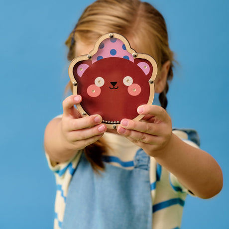 A young child with braided hair holds a cute bear mask over their face, sitting cross-legged. They wear a striped shirt and denim outfit, set against a blue background.