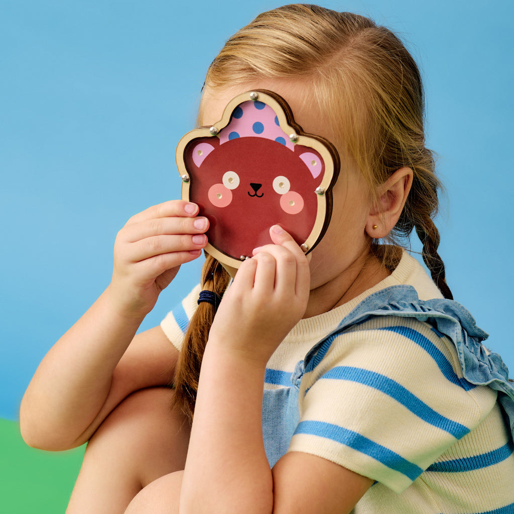 A young child with braided hair holds a cute bear mask over their face, sitting cross-legged. They wear a striped shirt and denim outfit, set against a blue background.