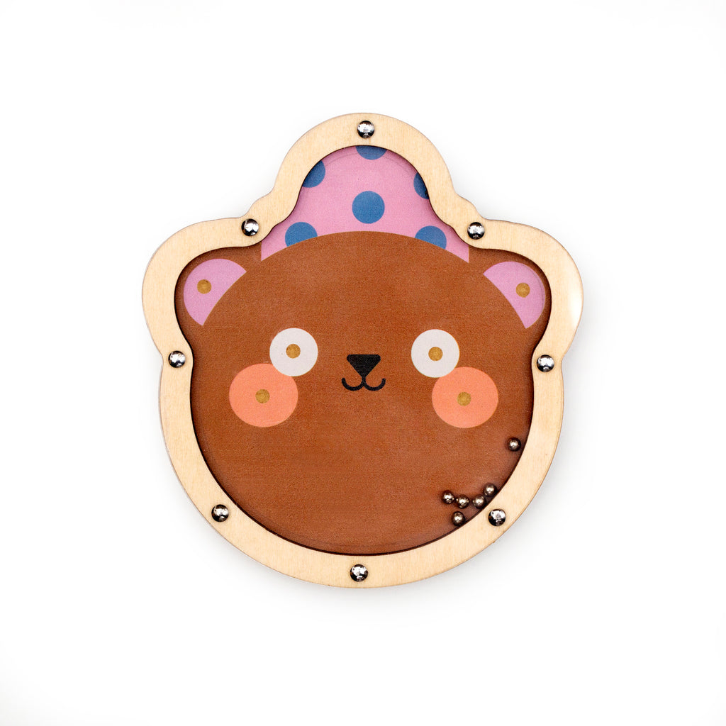 A cute, cartoonish bear face with a smile, framed by a wooden border. The bear has rosy cheeks and a pink hat with blue polka dots, evoking a playful mood.