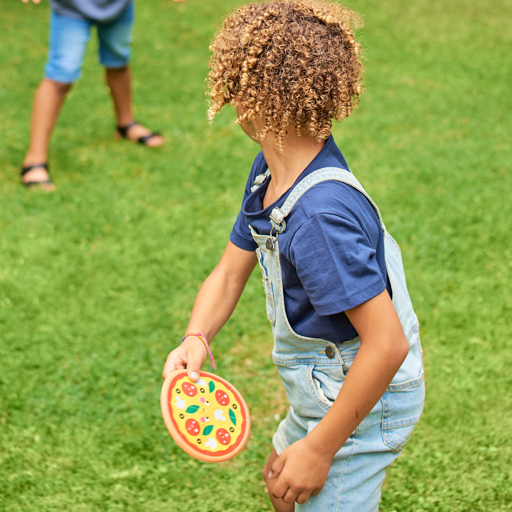 A child with curly hair, wearing a blue shirt and denim overalls, plays on grass, holding a round toy shaped like a colorful pizza, suggesting outdoor fun.