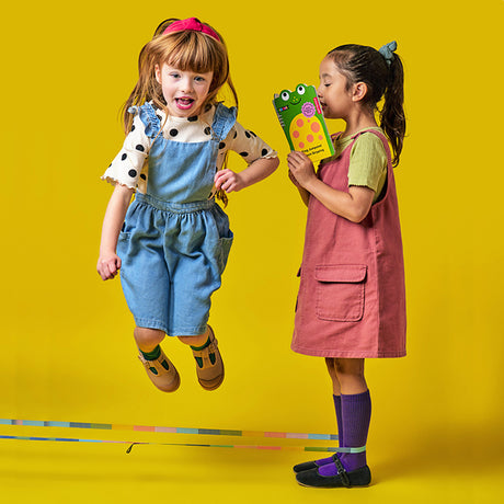 Two young girls against a bright yellow background. One jumps excitedly, wearing a dotted shirt and denim overalls. The other smiles, holding a colorful book.