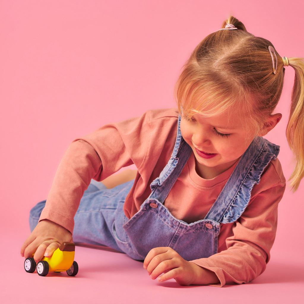 A smiling child with blonde pigtails plays with a yellow toy car on a pink background. She wears a peach shirt and denim overalls, conveying a cheerful mood.