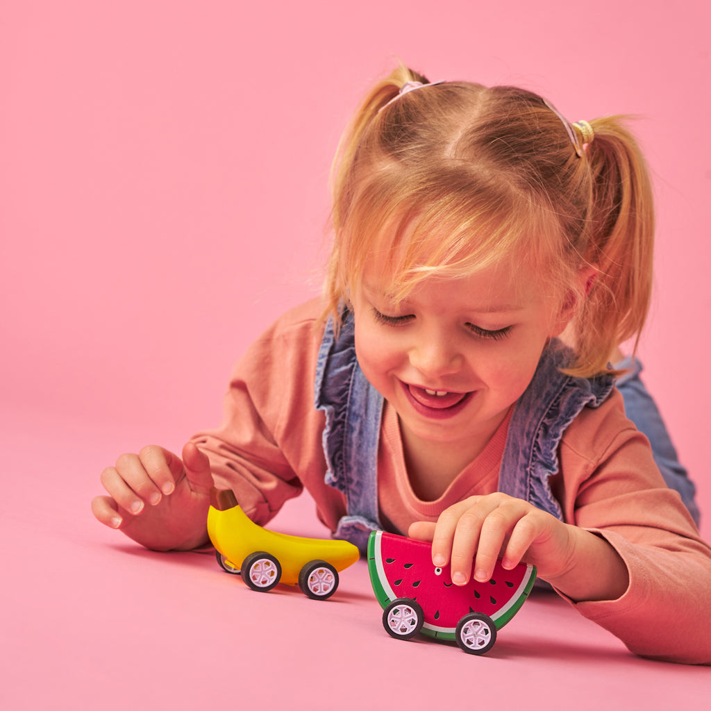 A cheerful child with blonde pigtails plays on a pink background, pushing toy cars shaped like a banana and a watermelon slice, conveying joy and playfulness.