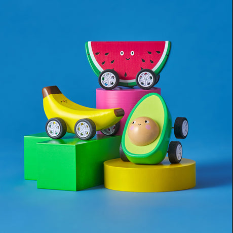 Toy cars shaped like an avocado, banana, and watermelon slice on colorful blocks against a blue background. The toys have painted smiling faces.