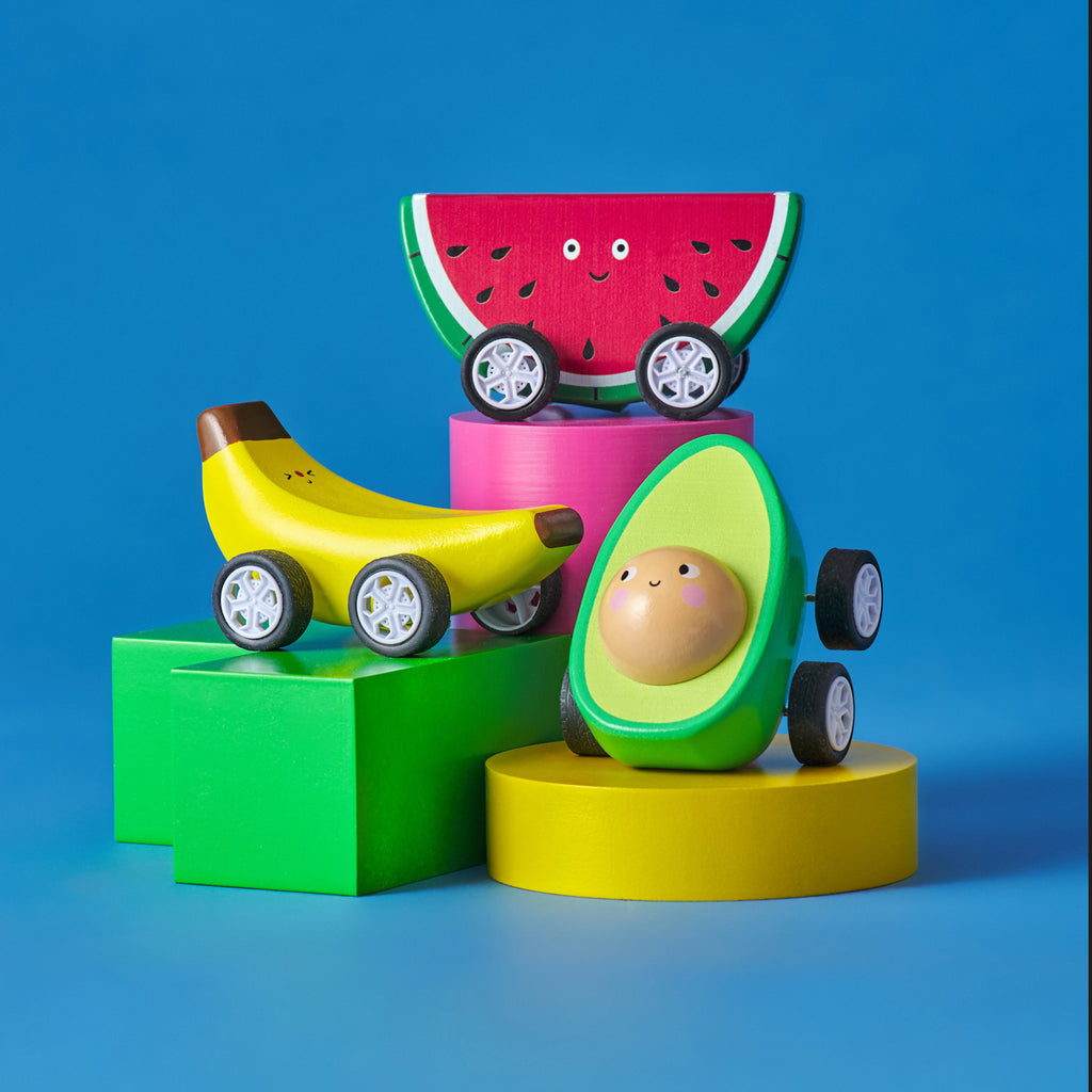 Toy cars shaped like an avocado, banana, and watermelon slice on colorful blocks against a blue background. The toys have painted smiling faces.