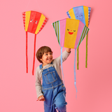 A child kneels joyfully on a pink background, holding a colorful kite with a smiling face. Two more vibrant kites hang above, creating a playful scene.